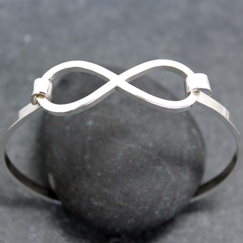 May include: A silver-colored bangle bracelet featuring an infinity symbol at the center. The bracelet has a simple, elegant design with a smooth, polished finish. The infinity symbol is connected to the bracelet's band with small rectangular connectors.