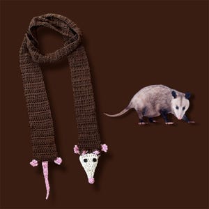 May include: A brown crochet scarf with a white and pink opossum head and tail design. A realistic opossum is next to the scarf on a brown background. The scarf is handmade and features a unique animal-themed design.