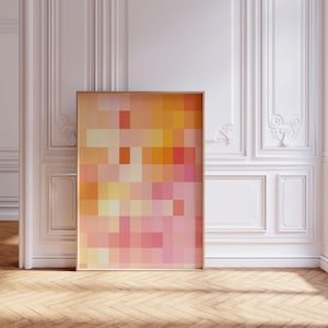 May include: A framed abstract art print featuring a pixelated design in shades of orange, yellow, and pink. The artwork is displayed against a white wall with decorative molding and a wooden floor.