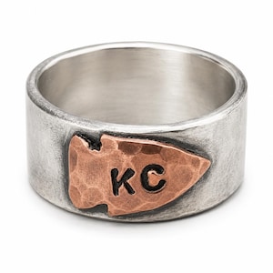 Op de afbeelding: Een brede zilveren ring met een gehamerde koperen pijlpunt als inleg aan de voorzijde. De koperen pijlpunt heeft een getextureerd oppervlak en is gestempeld met de zwarte letters KC. De zilveren ring heeft een geborstelde, matte afwerking.