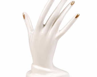 Op de afbeelding: Een witte, handvormige sieradenhouder van keramiek met een glanzende afwerking. De vingers zijn rechtop uitgestrekt en elke vingertop is voorzien van een metallic gouden accent dat doet denken aan gelakte vingernagels.