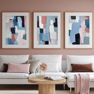 Pode incluir: Três pinturas abstratas em molduras de madeira, com formas geométricas em tons de azul, rosa e branco, penduradas em uma parede rosa claro acima de um sofá seccional branco com almofadas rosa e marrom.