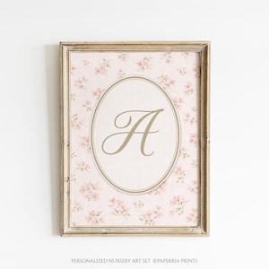 Puede incluir: Impresión floral enmarcada con fondo rosa y pequeñas flores rosas. Un óvalo central contiene la letra "A" en escritura dorada. El marco es dorado. El texto inferior dice "PERSONALIZED NURSERY ART SET ©PAPERIA PRINTS."