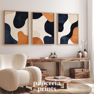 May include: Three framed abstract art prints with organic shapes in navy, tan, and orange hues. The artwork hangs above a wooden console table with a white armchair and a round coffee table in front. The prints are in wooden frames.