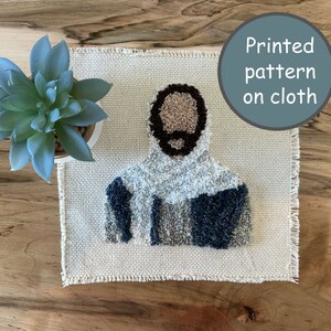 Puede incluir: Un paño beige con un patrón impreso de una persona con barba marrón y una prenda con capucha azul y gris. El texto "Patrón impreso en tela" está impreso en un círculo azul en la esquina superior derecha de la imagen.