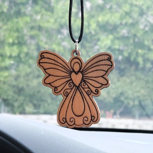 May include: A wooden angel car charm with a heart design hanging from a black string. The angel is intricately carved with detailed wings and a heart in the center of its chest.
