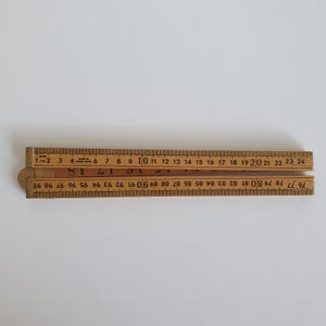 May include: A vintage wooden folding ruler, with inch and centimetre measurements. The ruler is made of light brown wood with black and gold markings. The ruler is partially open, displaying both sides with numbers.