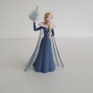 以下が含まれることがあります： 三つ編みのブロンドヘアのキャラクターのプラスチック製フィギュア。赤い斑点のある高い白い襟のダークブルーのロングドレスと、半透明の青いマントを着用し、小さく透明なギザギザの氷の結晶のような構造物を手に持っている。
