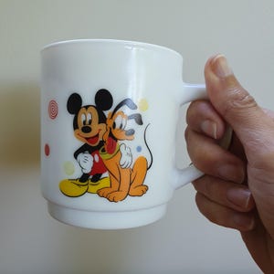 May include: White ceramic mug featuring Mickey Mouse and Pluto hugging. The mug has a handle and is decorated with colorful cartoon graphics. The design includes red, yellow, and black colors. The mug is held by a hand.