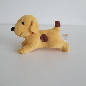 Op de afbeelding: Een pluche hondenspeeltje in een goudgele kleur met een donkerbruine vlek op de zijkant, een donkerbruine staartpunt en een zwarte neus. Het speeltje heeft een geborduurde mond en ogen, met een klein wit stoffen label aan de achterkant met tekst.