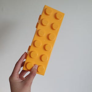 May include: A rectangular yellow plastic storage case shaped like a large building block. The top surface features ten raised circular studs, each embossed with the LEGO logo. The case has a hinged side design and a smooth, solid yellow finish.