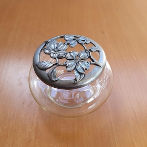 May include: A clear glass jar with a decorative metal lid. The lid features a floral design with detailed flowers and leaves. The jar is round and sits on a wooden surface. The lid is a silver colour.