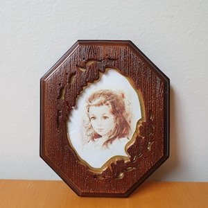 May include: An octagonal wooden picture frame with a portrait of a young girl. The frame has a dark brown finish with carved decorative elements. The portrait features a girl with long, curly hair and a soft, vintage aesthetic.