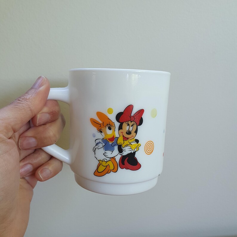 May include: White ceramic mug featuring Daisy Duck and Minnie Mouse. Daisy Duck is wearing a blue top and Minnie Mouse is wearing a yellow top. Both characters are smiling. The mug has a white handle and a white interior.