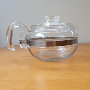 May include: A clear glass teapot with a rounded body, a curved handle, and a lid. A silver-coloured metal band encircles the middle of the teapot. The teapot is on a light brown wooden surface.