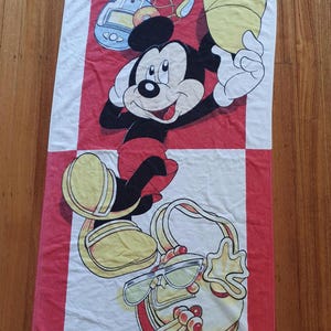 May include: A rectangular beach towel featuring Mickey Mouse in a playful pose. The towel has a red and white checkered design with Mickey Mouse holding a yellow object. The towel also features a purse and sunglasses.