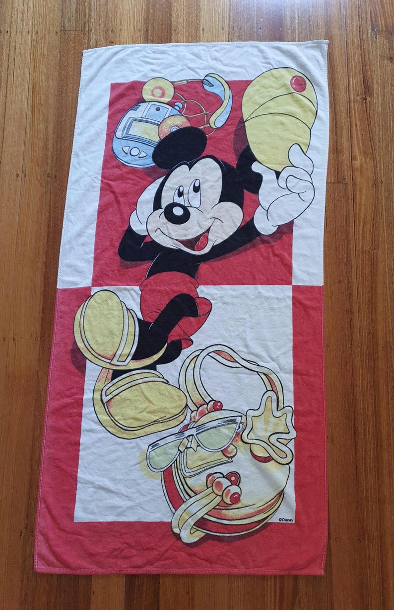 May include: A rectangular beach towel featuring Mickey Mouse in a playful pose. The towel has a red and white checkered design with Mickey Mouse holding a yellow object. The towel also features a purse and sunglasses.