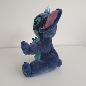 May include: A plush toy of the character Stitch from the animated film Lilo & Stitch. The toy is primarily blue with large ears, a tuft of hair, and large eyes. The plush is sitting with its arms outstretched.