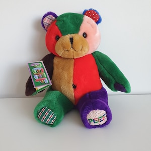 May include: A plush teddy bear made of multi-coloured fabric patches in red, green, brown, purple, and pink. The bear features a tag with the text PEEF attached to its arm and the word PEEF embroidered on its purple foot pad with a polka dot pattern.