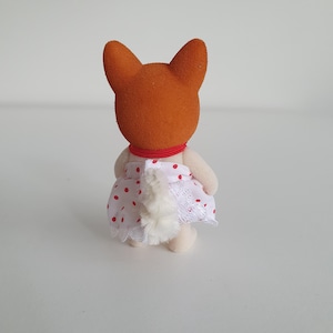 May include: A small, orange-headed fox figurine, viewed from the back, wearing a white dress with red polka dots and lace trim. The figurine has a red collar and a fluffy white tail. The toy is on a white surface.