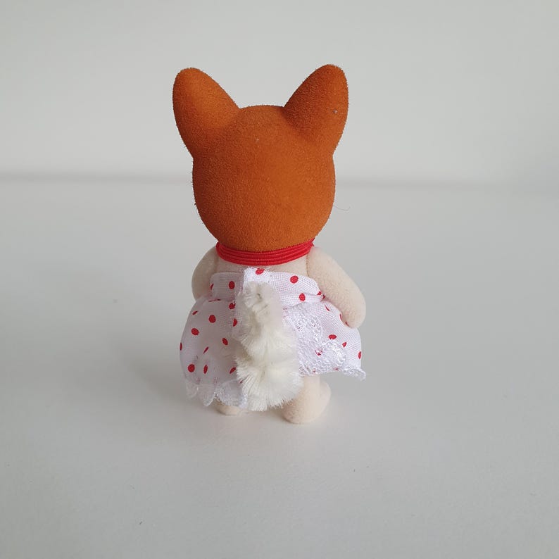 May include: A small, orange-headed fox figurine, viewed from the back, wearing a white dress with red polka dots and lace trim. The figurine has a red collar and a fluffy white tail. The toy is on a white surface.