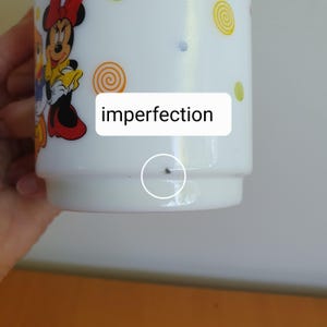 May include: White ceramic mug featuring a Minnie Mouse cartoon design with red, yellow, and black accents. The mug has a spiral and dot pattern. The word "imperfection" is written on the mug.
