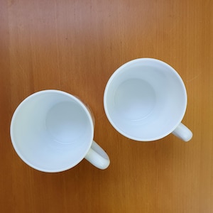 May include: Two white ceramic coffee mugs with curved handles are positioned on a wooden surface. The mugs are empty, and the interior is visible. The mugs are positioned at an angle, offering a top-down view.