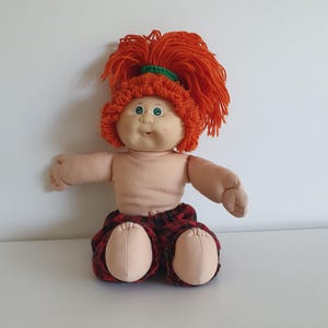 以下が含まれることがあります： 柔らかい肌色の体、緑色の目、笑顔の表情をしたヴィンテージ人形。人形は明るいオレンジ色の糸の髪をしており、赤と黒のチェック柄のショートパンツを履いています。人形は腕を広げて座っています。