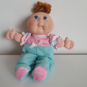 May include: A doll with a round face, blue eyes, freckles, and reddish-brown hair tied in a top knot. The doll wears a pink top with floral ruffled sleeves and a white bow, paired with teal trousers featuring pink fabric on the feet.