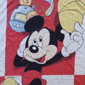 May include: A white towel featuring a smiling Mickey Mouse character in a red and black design. Mickey is wearing red shorts and holding a yellow hat. The background includes a red and white checkered pattern and a retro music player.