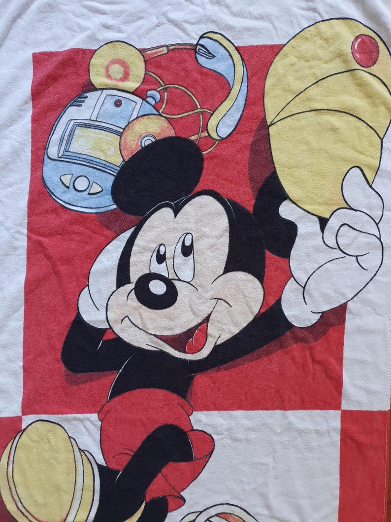 May include: A white towel featuring a smiling Mickey Mouse character in a red and black design. Mickey is wearing red shorts and holding a yellow hat. The background includes a red and white checkered pattern and a retro music player.