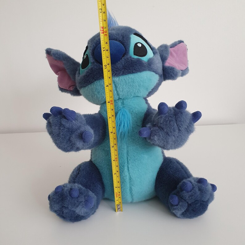 May include: A plush Stitch doll, primarily blue with turquoise accents, and pink inner ears. The doll is approximately 13 inches tall. The character has large black eyes and a tuft of blue fur on its chest. The doll is sitting on a white surface.