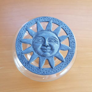 May include: A clear glass jar with a decorative lid featuring a smiling sun design. The lid is a dark blue-grey colour with a textured surface and sun rays. The jar sits on a light brown wooden surface.