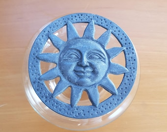 May include: A clear glass jar with a decorative lid featuring a smiling sun design. The lid is a dark blue-gray color with a textured surface and sun rays. The jar sits on a light brown wooden surface.