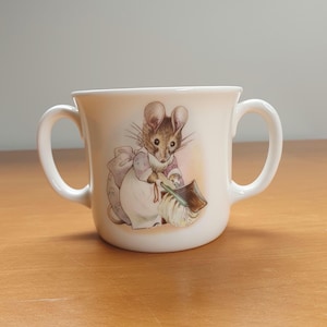 May include: A white ceramic mug with two handles features a Beatrix Potter illustration of a mouse holding a broom and dustpan. The mouse is wearing a pink dress and apron.