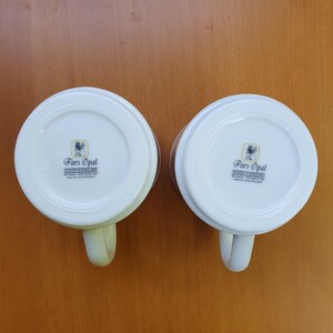 May include: Two white ceramic mugs with handles, featuring the "Pars Opal DESIGN IN ENGLAND" logo on the bottom. The mugs are set against a light brown background, showcasing their clean design and classic shape.