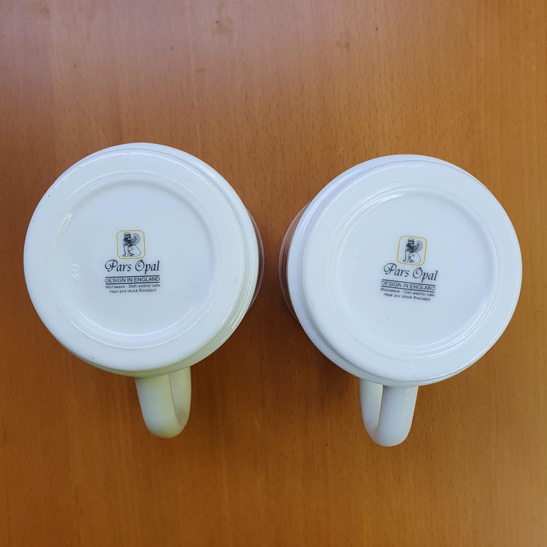 May include: Two white ceramic mugs with handles, featuring the "Pars Opal DESIGN IN ENGLAND" logo on the bottom. The mugs are set against a light brown background, showcasing their clean design and classic shape.