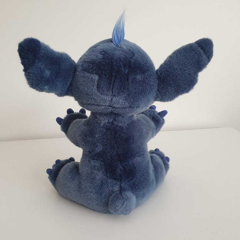 May include: A plush toy of the character Stitch from Lilo & Stitch, viewed from the back. The toy is a shade of blue with large ears, a tuft of hair, and small claws. It is sitting on a white surface.