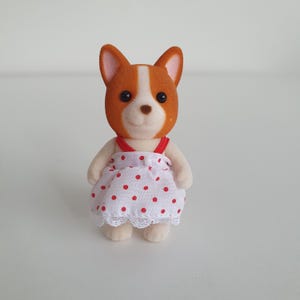 May include: A small, plush dog figurine with brown and white fur, wearing a white dress with red polka dots and a red strap. The dress has a lace trim. The dog has black eyes and a smiling expression.