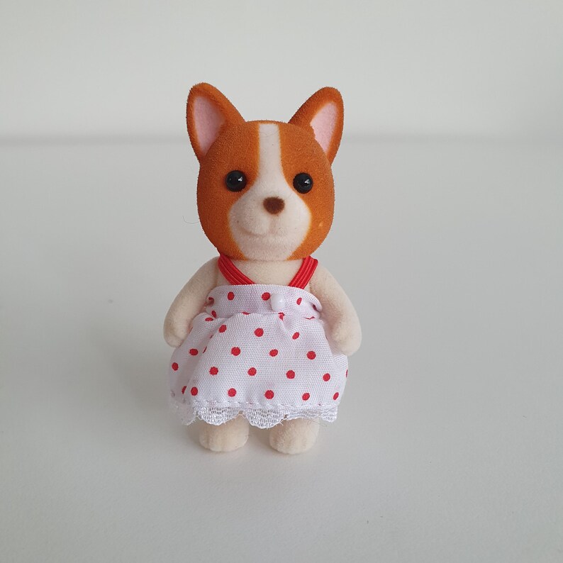 May include: A small, plush dog figurine with brown and white fur, wearing a white dress with red polka dots and a red strap. The dress has a lace trim. The dog has black eyes and a smiling expression.