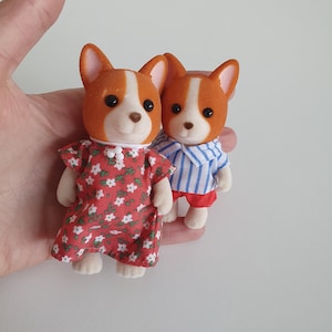 May include: Two miniature dog figurines. One wears a red floral dress, the other a blue and white striped shirt and red shorts. Both have orange and white fur, black eyes, and are held in a hand.