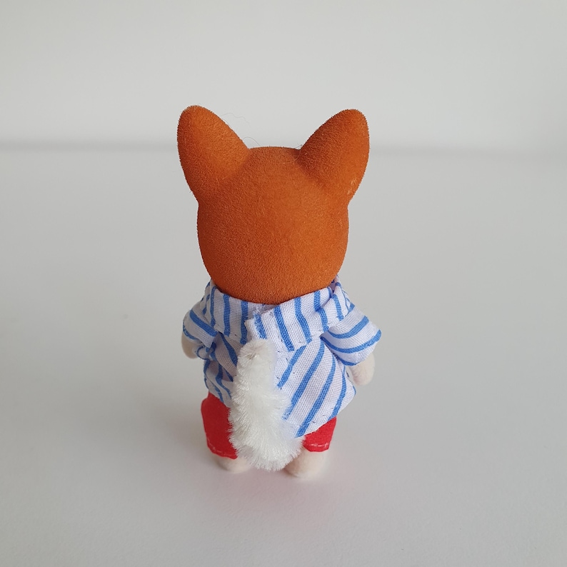 May include: A small, orange fox figurine, viewed from the back, wearing a blue and white striped shirt and red pants. The fox has a fluffy white tail. The figurine is a toy.