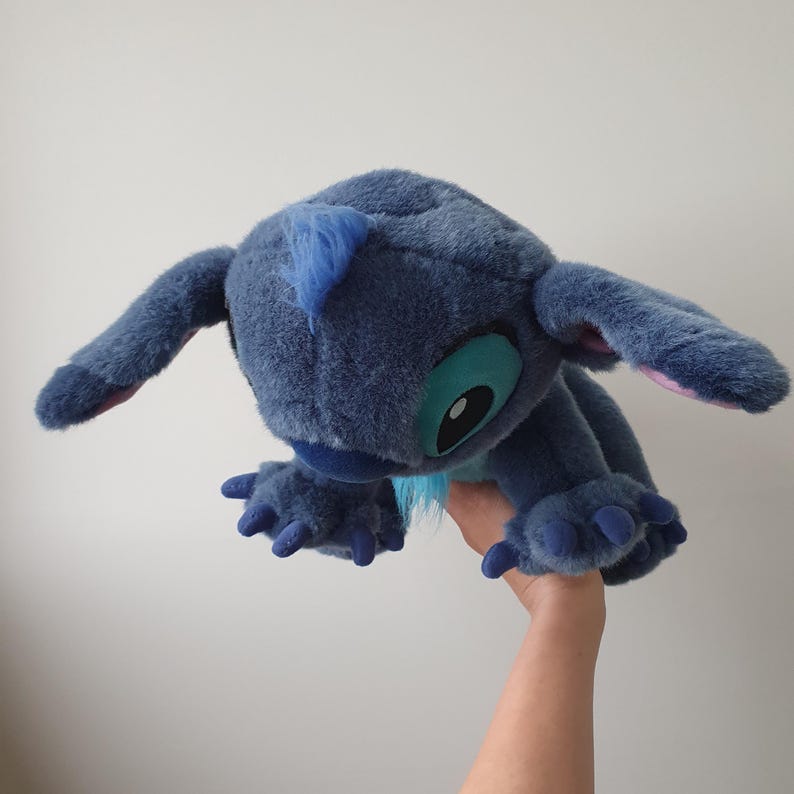 May include: A plush toy of the character Stitch from the animated film Lilo & Stitch. The toy is primarily blue with large, expressive eyes, long ears, and a tuft of blue hair. The toy's claws and inner ears are pink.