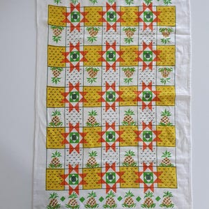 Puede incluir: Un paño de cocina rectangular blanco con un patrón de cuadrícula repetitivo de ilustraciones de piñas amarillas y motivos de estrellas geométricas naranjas y verdes. El diseño está dispuesto en filas alternas.