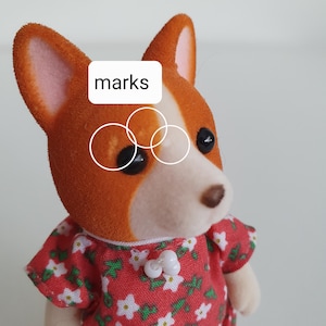 May include: A close-up of a corgi dog figurine with orange and white fur, wearing a red floral dress. The figurine has black eyes and a brown nose. The word "marks" is in a white rectangle above the dog's eye.