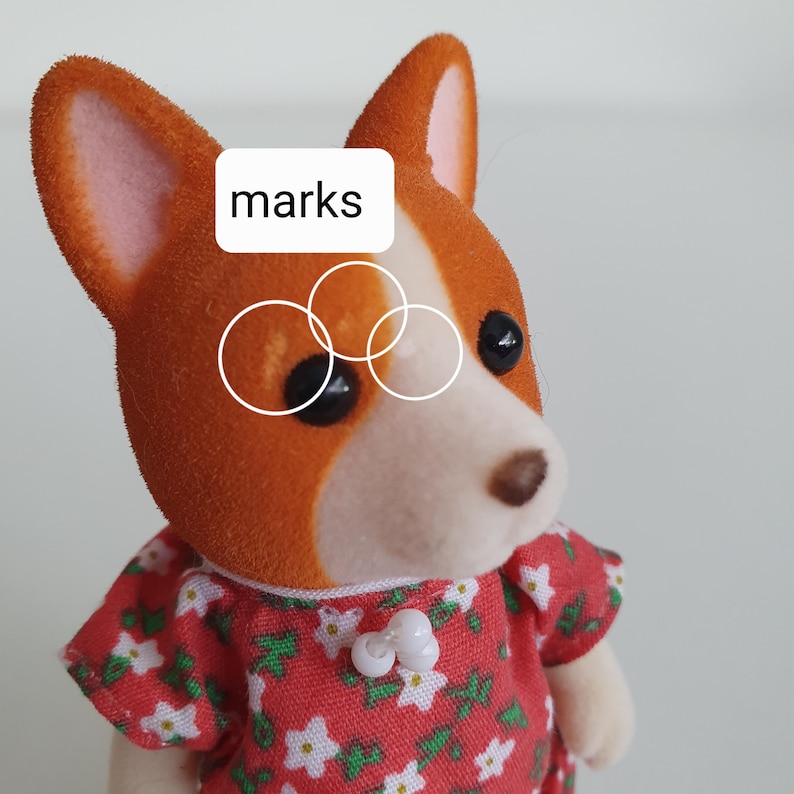 May include: A close-up of a corgi dog figurine with orange and white fur, wearing a red floral dress. The figurine has black eyes and a brown nose. The word "marks" is in a white rectangle above the dog's eye.
