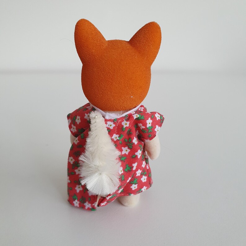May include: A small toy fox figure with orange fur and a red dress with white floral patterns. The figure has a white fluffy tail and is viewed from the back.