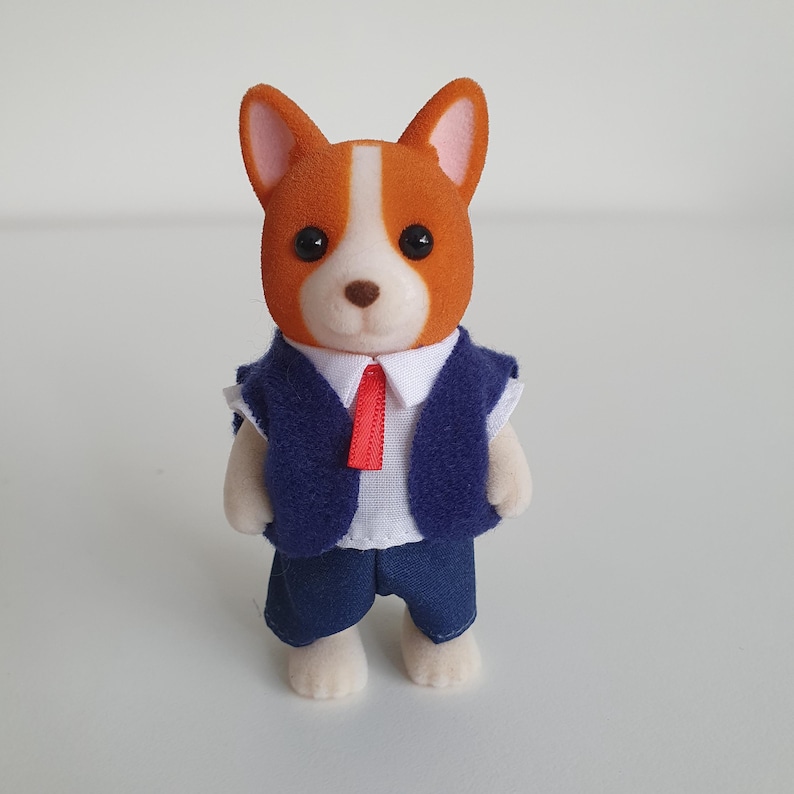 May include: A small, orange and white dog figurine wearing a blue vest, shorts, and a red tie. The dog has black eyes and a brown nose. The figurine is standing upright.