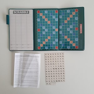 May include: A folding Scrabble board game set featuring a dark green case with a score sheet on the left panel, a grid board in the center and right panels, a printed rules sheet, and a sheet of beige letter tiles with black letters and numbers.