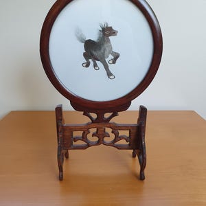 May include: A round wooden frame with a dark brown horse embroidered on a white background. The frame is on a wooden stand.